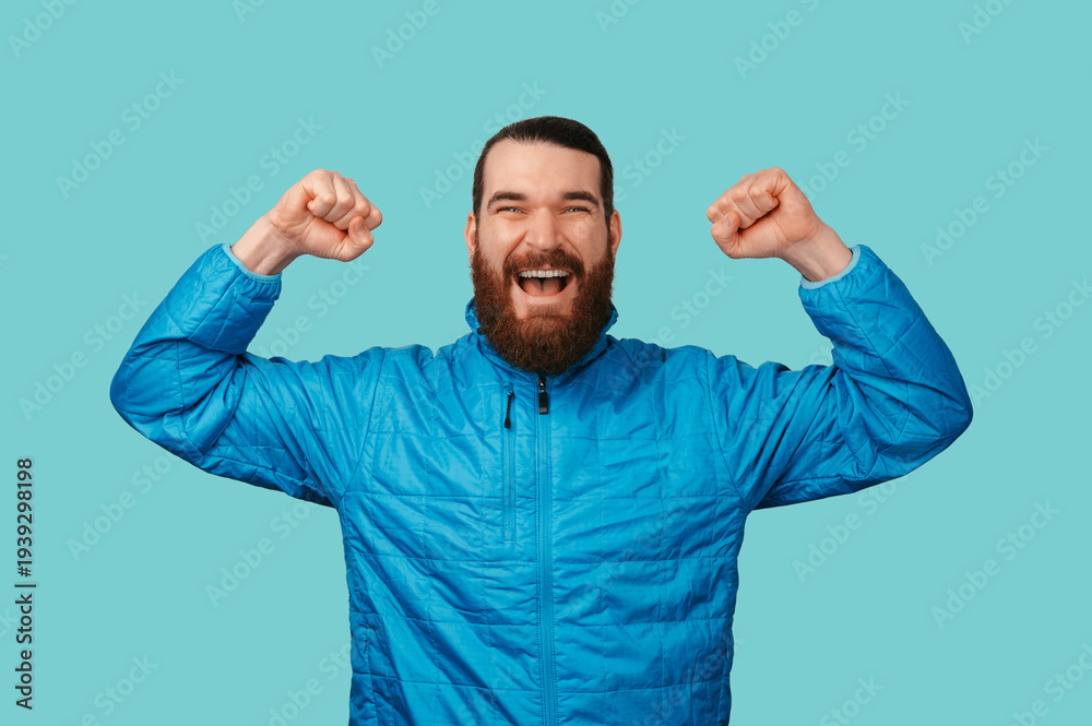 Fototapeta premium Triumphant Caucasian Man With Raised Fists, Joyful Expression Against Turquoise Backdrop Wearing Blue Jacket,