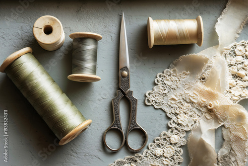 Vintage ornate dressmaking scissors with wooden thread spools and delicate cream lace trim flat lay on gray background.