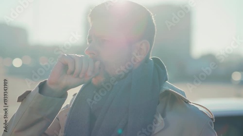 Man lingers outdoors coughing and adjusting his scarf, Man in parking lot coughs and modifies his scarf while outdoors