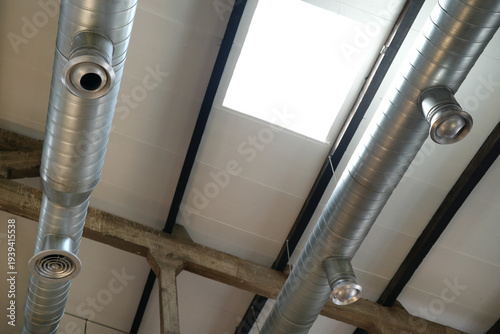 A low-angle view of two parallel silver metal spiral ducts with circular air diffusers installed on a ceiling with a skylight in an industrial or commercial building.