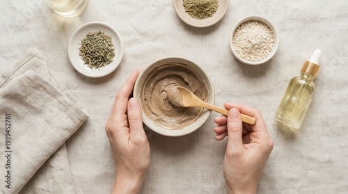 Organic skincare ingredients natural clay mask preparation on linen table, female hand mixing powder with wooden spoon near glass serum bottle, calm minimal spa ritual at home