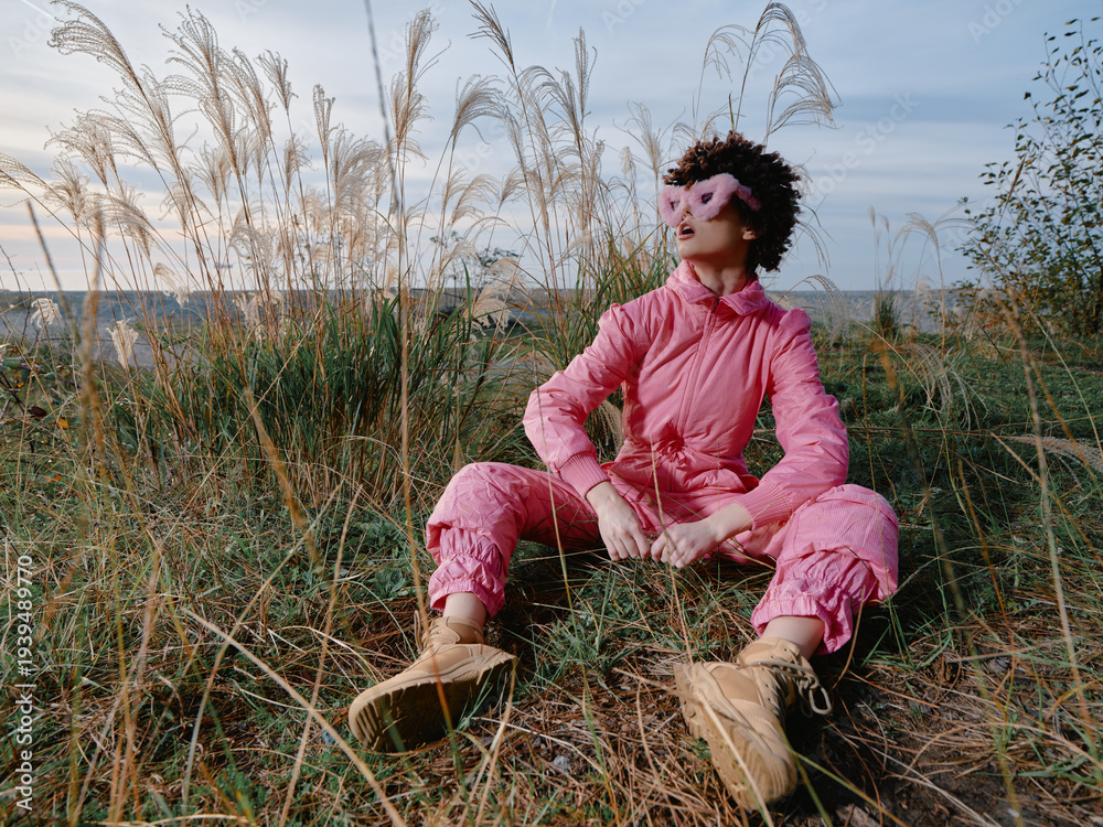 Fototapeta premium Outdoor person wearing pink fashion sits among tall grasses in a field, relaxed pose with boots, wide sky, natural setting, calm mood, candid lifestyle scene for travel or editorial use