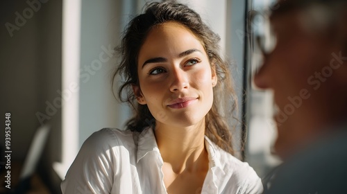 A young woman in a white shirt looking thoughtful and relaxed while engaged in a conversation near a sunlit window