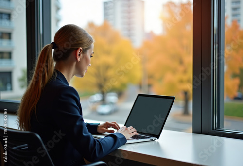 Business person working on laptop by window, autumn city view
