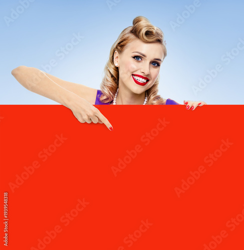 Expressive smile woman wear pinup dress show point finger red blank banner, mockup free text space. Blonde pin up girl advertise, isolated blue background. Retro vintage sales ad image