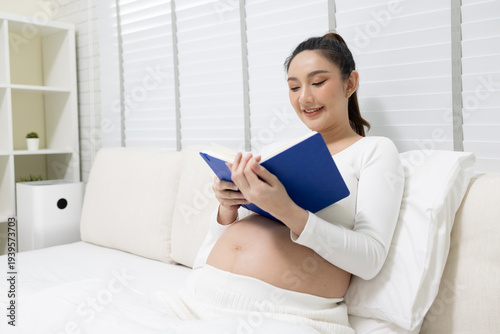 Happy pregnant Asian woman sitting on a white bed and reading a blue book. Ideal for prenatal care, motherhood lifestyle, pregnancy health, home relaxation, and family preparation concepts.