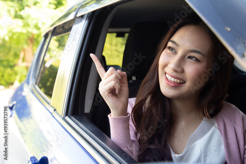 A happy Asian woman is sitting in a blue car and pointing her finger out of the window. Great for road trip concepts, new driver insurance, and summer travel
