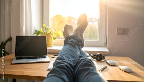Relaxed person with legs stretched out under a wooden desk in a bright home office while sunlight streams through the window
