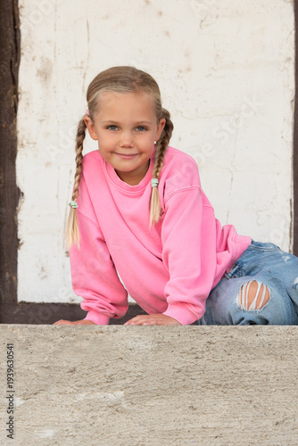 little smiling girl with braided pigtails, pink jumper, sitting sideways in front of white wall and stone in front