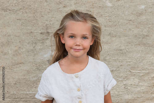 portrait of little friendly smiling girl with long blond hair in white dress in front of stone wall - natural look