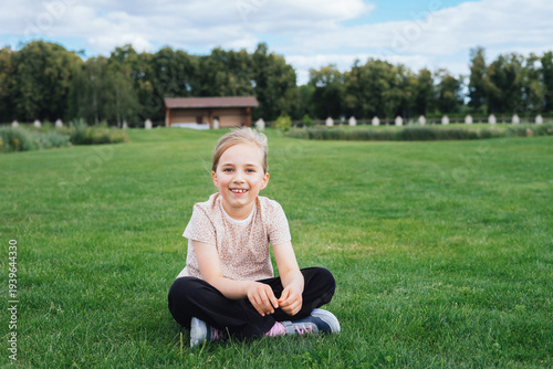 Smiling young girl sitting cross-legged on a lush green lawn at the park, open space green background