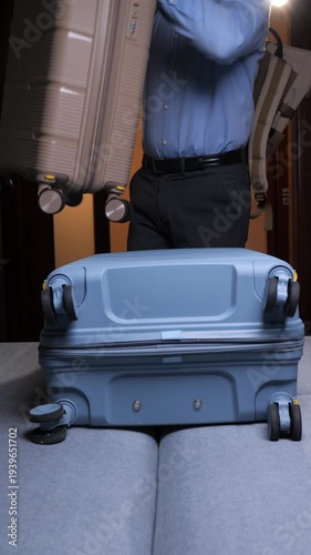 Vertical video man standing by an open blue hardshell suitcase, wearing a blue shirt and backpack, getting ready for a trip and organizing belongings in a room