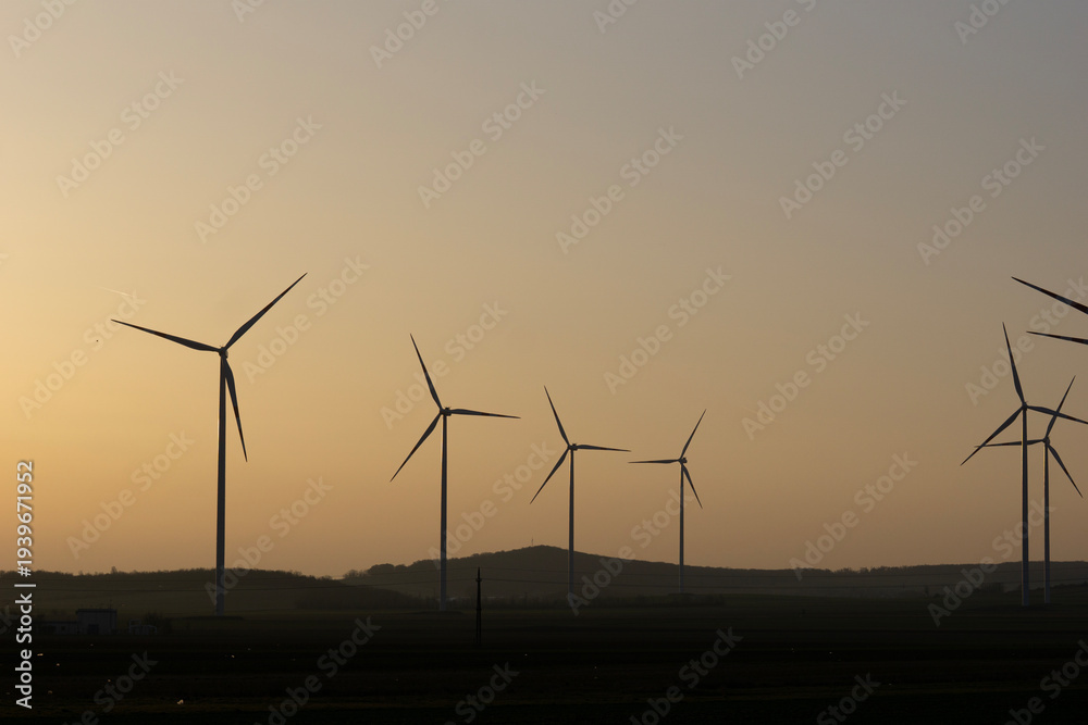 Naklejka premium a wide shot of several large wind turbines silhouetted against a hazy sunset sky over a dark rural field in a calm evening atmosphere