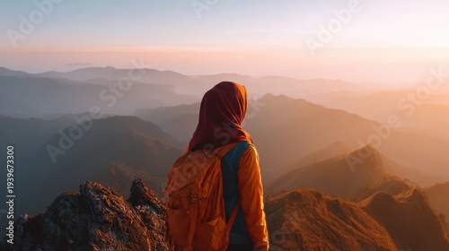 Muslim woman in orange hijab sitting on rocky mountain peak gazing at misty mountain landscape