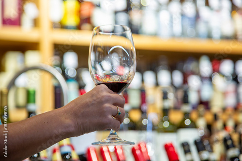 Wine tasting. A woman's hand holds a glass of red wine against the backdrop of a store window displaying bottles of wine.