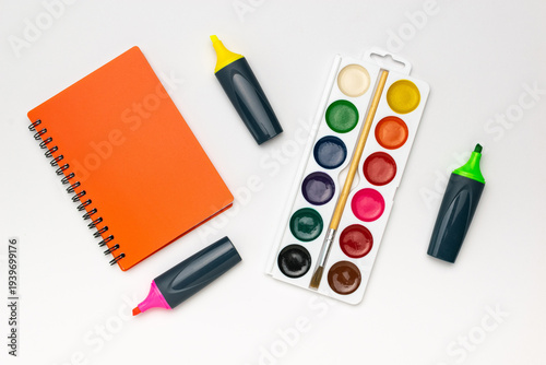 Creative Workspace: Orange Notebook, Watercolor Palette, Highlighter Pens on White Background
