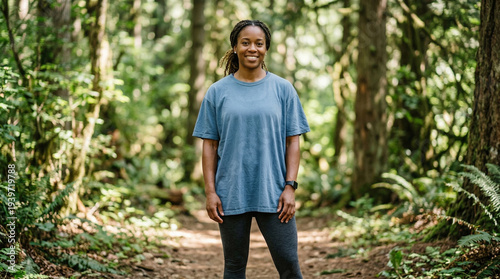 A woman stands on a dirt trail surrounded by trees in a forest. She is wearing a blue t-shirt for mockup.