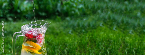 Refreshing summer lemonade with strawberries and citrus. Pouring fresh water into a glass jug with fruit and mint on a green grass background. Wide banner with copy space