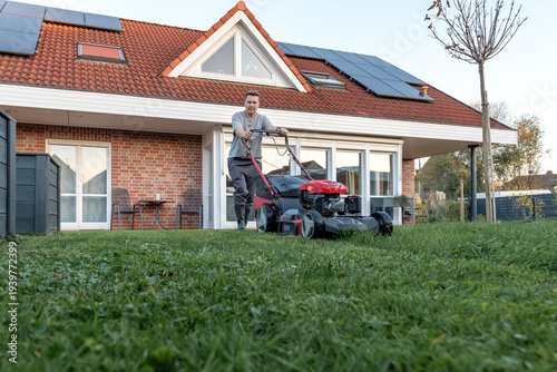 Man mowing narrow backyard lawn beside modern metal fence and garden plants.