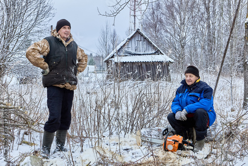 Standing in snowy forest clearing, lumberjack rests during break while partner sits beside chainsaw near small wooden shed.