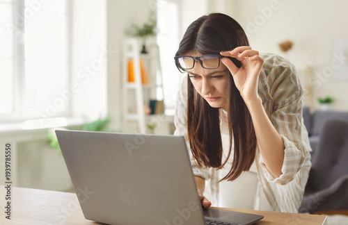 Canvas Print Young woman leaning toward laptop and lifting glasses while struggling to read text