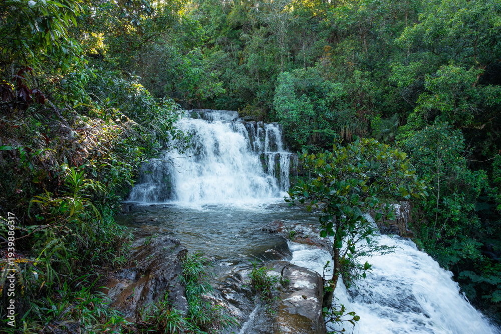 Obraz premium Waterfall cascading over multi-tiered rocky formations surrounded by lush green tropical forest, expressing the power of water, natural beauty, and untouched wilderness