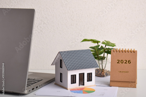 Miniature house with laptop and 2026 desk calendar on workspace. Real estate planning, property investment strategy, housing market outlook, and home buying financial management concept.