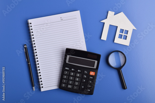 Real estate concept. White paper house, calculator, blank notebook, pen, and magnifying glass on blue background. Perfect for mortgage planning, property appraisal, and home budgeting.