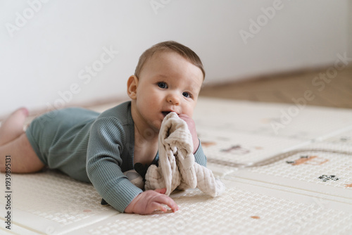 Adorable baby lying on tummy on a soft play mat and playing with a sensory toy. The infant is focused and curious while exploring the toy, supporting early learning and motor skill development.