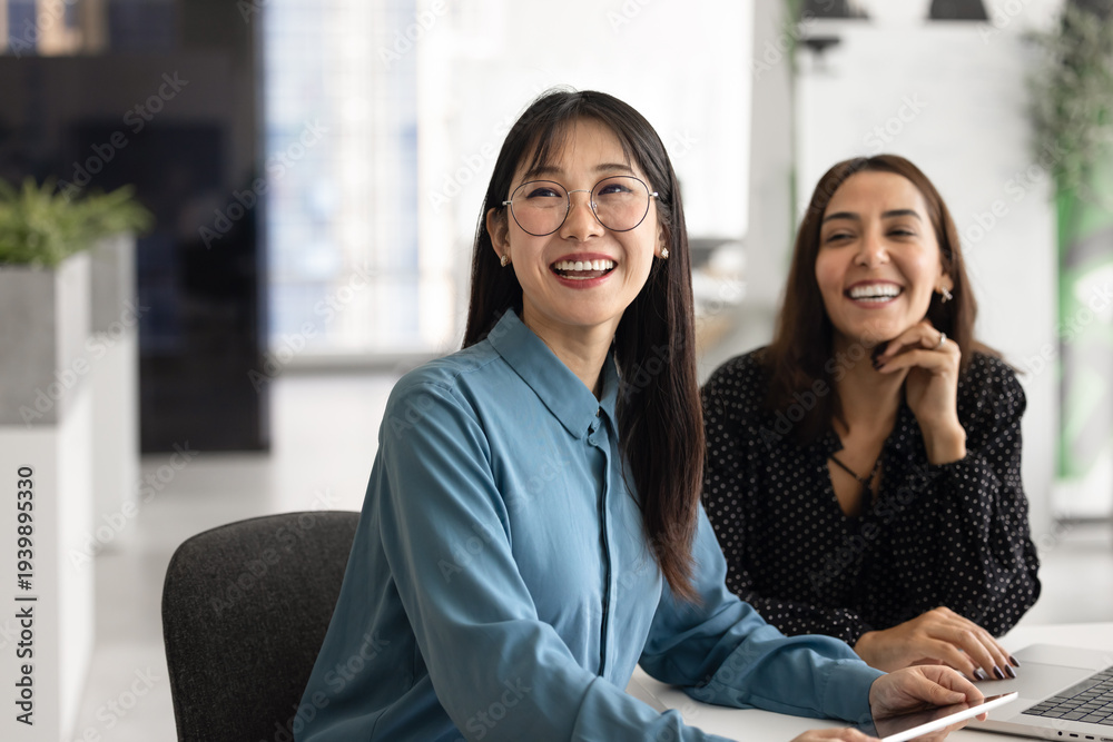 Obraz premium Happy young Asian business professional woman in glasses sitting at office table, working with positive Middle Eastern female coworker together, looking away, smiling, laughing