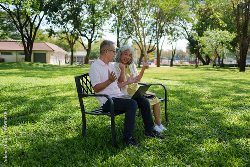 Cheerful senior couple sitting on bench in green park holding laptop computer talking with joyful emotional expression during outdoor retirement