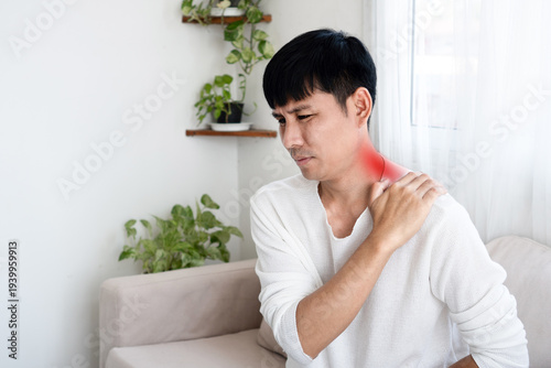 Slika na platnu Asian man suffering from severe neck and shoulder pain at home, touching inflamed area with red highlight, feeling exhausted and stressed from office syndrome or muscle strain