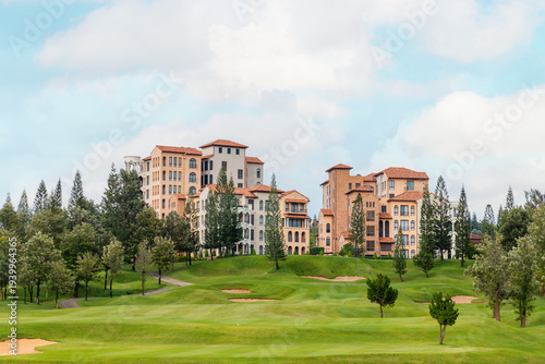 Luxury Mediterranean style buildings stand on a hill overlooking a lush green golf course fairway with sand traps and pine trees under a cloudy blue sky. High-end real estate and vacation resort.