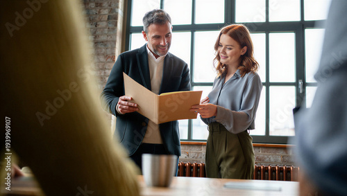 Senior art director and young designer experiencing quiet pride while reviewing physical design proofs during a crowded creative agency meeting.