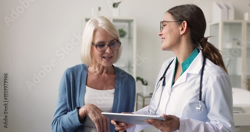 Female doctor holds digital tablet, explaining telehealth application use, sharing medical information, clarifying checkup results to middle age woman patient during consultation. Healthcare services