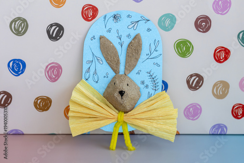 A charming handmade Easter bunny craft, fashioned from recycled materials and adorned with a bright yellow pleated bow. It's set against a light blue card with delicate floral doodles.