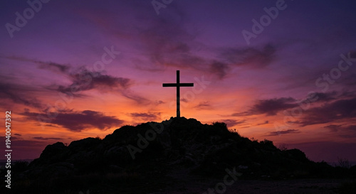 A silhouette of a cross stands atop a rocky hill against a dramatic sunset sky. The backdrop features a vivid gradient of colors, from deep purple to fiery orange, with scattered clouds adding texture