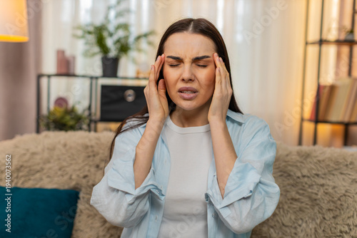 Canvas Print Young woman at home sitting on sofa holding head in hands, experiencing severe headache discomfort