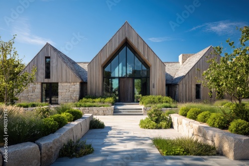 A contemporary home featuring a stunning facade of wood and stone is beautifully nestled within a lush green landscape, all illuminated by bright daylight beneath a clear blue sky
