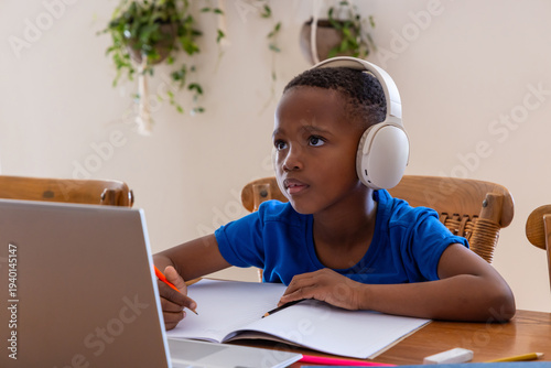 Male child wearing white headphones blue shirt studying at home table laptop notebook orange pencil
