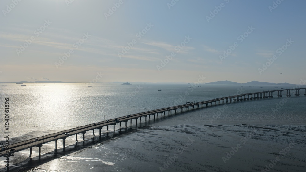 Obraz premium Aerial drone photography of the long curved path of Incheon Bridge stretching over the shimmering sea under a soft blue sky at golden hour in South Korea.