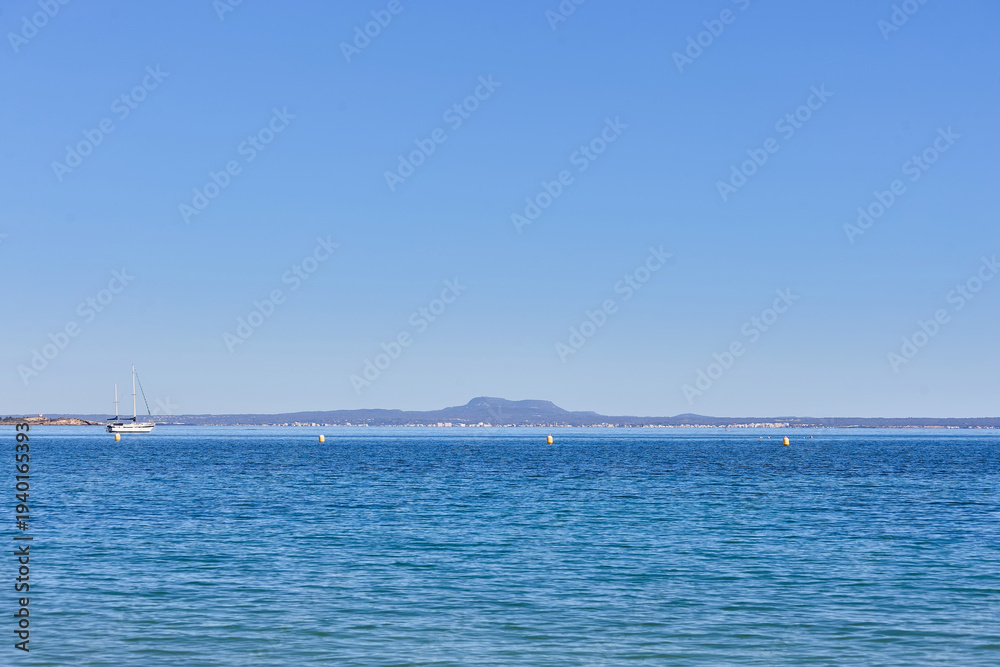Fototapeta premium Open view across calm Mediterranean water toward distant mountains on the horizon near the coast of Mallorca, Spain, with a small sailboat and navigation buoys visible offshore.