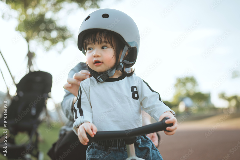 Naklejka premium Close-up of a cute Asian toddler girl wearing a safety helmet while learning to ride a balance bike at a public park during a sunny afternoon, representing early childhood development, outdoor wellnes