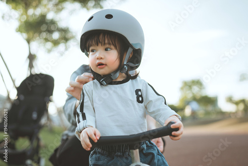 Close-up of a cute Asian toddler girl wearing a safety helmet while learning to ride a balance bike at a public park during a sunny afternoon, representing early childhood development, outdoor wellnes