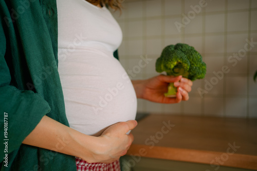 Pregnant woman in a home kitchen gently holding her baby bump with one hand and offering fresh broccoli with the other, symbolizing prenatal healthy eating and mindful nutrition for mom and baby