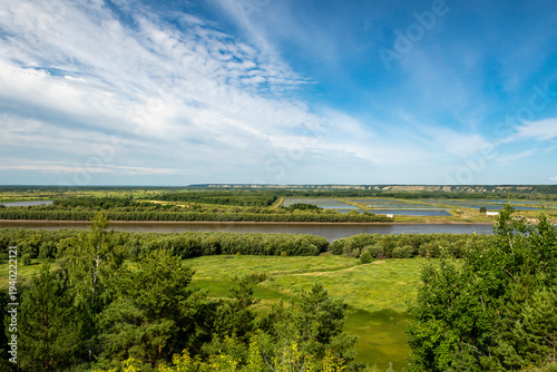 A river flows through a grassy green plain, and hills are visible in the background. The sky is overcast, giving the scene a serene atmosphere.