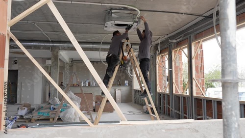 Two professional hvac technicians standing on ladders and installing a ceiling mounted air conditioner unit during the renovation of a commercial building, focusing on climate control