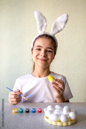 Smiling girl wearing bunny ears painting a yellow Easter egg at table, feeling playful and creative