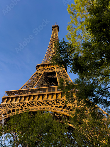 A stunning view of the Eiffel Tower rises above lush green trees under a clear blue sky. Visitors admire the iconic landmark while enjoying the pleasant weather in spring.