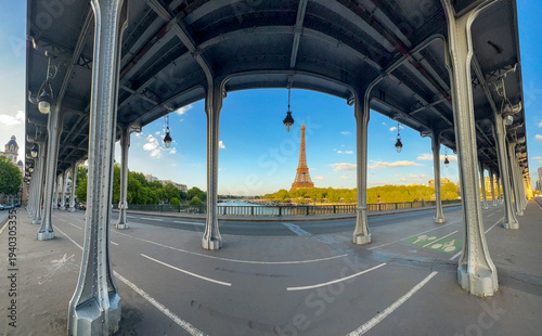 A clear blue sky frames the iconic Eiffel Tower, observed from under the Bir-Hakem Bridge. The scene captures the beauty of Paris along the Seine River on a tranquil afternoon.